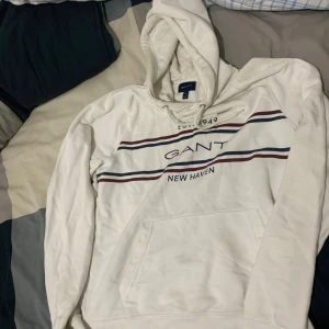 Vit hoodie från GANT med tryck - Vit hoodie från GANT med marinblå och vinröda ränder samt tryck 'GANT New Haven' på bröstet. Klassisk känguruficka, snörning i huvan och mjukt material. Perfekt för en chill och stilren look.