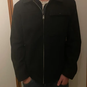 Svart ulljacka från Jack & Jones - Stilren svart ulljacka från Jack & Jones med klassisk krage, rak passform och dragkedja framtill. Jackan har en bröstficka med lock och långa ärmar. Perfekt för dig som vill ha en clean look med lite edge.