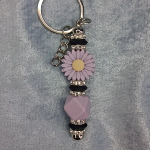 Lila blommig nyckelring - Handgjord nyckelring skapad med silikonpärlor i svart,lila, en stor lila tusensköna samt glittriga mellandelar. Perfekt som accessoar eller nyckelring för att ge din stil en personlig touch.