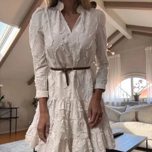 Vit kortklänning med strukturmönster - Supersöt vit kortklänning från H&M med lång ärm och volangkjol. Klänningen har ett unikt strukturmönster, v-ringning och är endast STYLAT med brunt bälte i midjan, YSL skärpet ingår inte. Perfekt för dig som gillar romantisk och trendig stil. Materialet känns lätt och luftigt, passar perfekt till ljusa dagar.