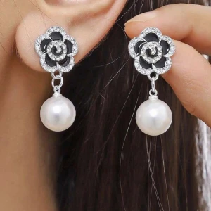 Svarta blomörhängen med pärlor - Unika örhängen med svart blomformad topp dekorerad med glittrande stenar och en stor vit pärla som hänger ned. Blommorna har silverkant och ger en lyxig vibe. Perfekt för dig som vill sticka ut med ett coolt och elegant smycke.