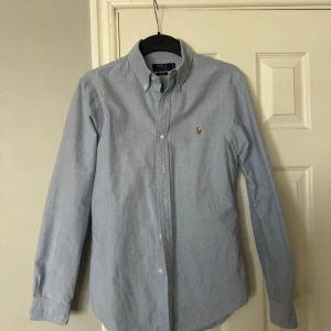 Blå skjorta från Polo Ralph Lauren - Klassisk ljusblå skjorta från Polo Ralph Lauren i slim fit-modell. Skjortan har button down-krage, långa ärmar och broderad logga på bröstet. Tillverkad i mjuk bomull och stängs med vita knappar framtill.