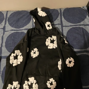 Svart hoodie från Denim Tears med blommor - Svart hoodie från Denim Tears med stora vita blommönster över hela tröjan. Klassisk huva och känguruficka framtill. Perfekt för dig som vill sticka ut med en streetwear vibe. Mjuk insida och snygg passform.