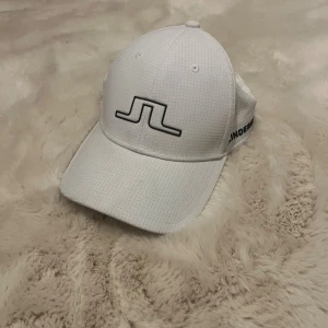 Vit keps från J.Lindeberg - Snygg vit (golf)/keps från J.Lindeberg med svart logotyp framtill och diskret rutmönster i materialet. Kepsen har böjd skärm och justerbar baksida. 