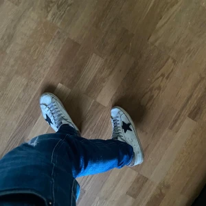 Golden Goose vita sneakers med svart stjärna - Golden Goose sneakers i vitt skinn med ikonisk svart stjärna på sidan och beige detaljer vid hälen. Skorna har snörning med GGDB-tryck och en klassisk rund tå. Perfekta för dig som gillar en avslappnad och trendig streetstyle.