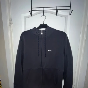 Svart hoodie med dragkedja från BOSS - Svart hoodie från BOSS med vit logga på bröstet och vita ribbade detaljer på ärmar och rygg. Tröjan har huva med snörning, hel dragkedja och känguruficka framtill. Perfekt för en chill och sportig look.