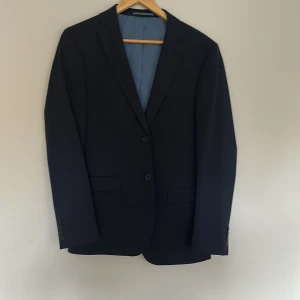 Mörkblå kavaj Dressmann slim fit storlek 48 - Snygg mörkblå kavaj från Dressmann i slim fit-modell. Kavajen har två knappar framtill, klassiska fickor och ljusblått innerfoder med röda detaljer. Perfekt för dig som vill ha en stilren och modern look. Kavajen är använd 1 gång.
