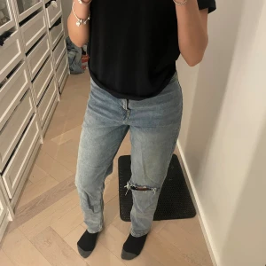 Blå jeans från Divided med hål - Säljer ett par blå jeans från H&M i rak modell med ett stort hål och slitning på ena knät. Jeansen har klassisk femficksdesign, normal passform och är tillverkade i bomull. Perfekta för en avslappnad och trendig look.