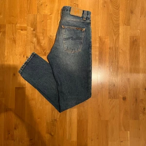Blå jeans från Nudie Jeans  - Säljer ett par klassiska och riktigt fräscha jeans från Nudie Jeans Co i storlek w28. Jeansen har raka ben, snygga kontrastsömmar och ikoniska bakfickor med vågig brodyr. Tillverkade i bomull med en autentisk tvättad look och orange detaljer. Byxorna är i excellent skick och säljs enbart för fel storlek!! Jag skickar samma dag eller senast dagen efter! Om någon vill ha mer bilder så är det bara att hojta till!!