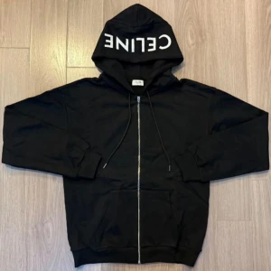 Celine hoodie (helt ny+gratis frakt) - Svart hoodie från Celine med vit logga på huvan, hel dragkedja framtill och snörning vid huvan. Tröjan har känguruficka och ribbade muddar. Perfekt för en chill och stilren look.