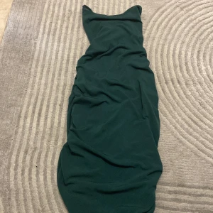 Mörkgrön långklänning med snörning - Säljer en mörkgrön långklänning med snygg snörning i ryggen och rynkade detaljer längs sidorna. Skronsh dör bak vid rumpan.Klänningen har en figurnära passform och är tillverkad i ett mjukt, stretchigt material. Perfekt för dig som vill sticka ut med en unik och trendig look.