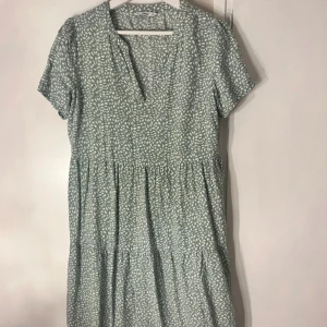 Grön kortklänning från Only - Söt grön kortklänning från Only med vitt bladmönster, v-ringad hals och korta ärmar. Klänningen har en avslappnad passform med rynkade detaljer i midjan och är tillverkad i mjuk bomull. Perfekt för dig som gillar en chill och fräsch stil.