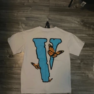 Vit Vlone t-shirt med fjärilstryck - Säljer en vit t-shirt från Vlone i storlek M med stor blå fjäril och orange V på framsidan, samt ett stort blått V med orange fjärilar på ryggen. T-shirten har rund hals och korta ärmar, tillverkad i mjuk bomull. Perfekt statement-plagg för dig som gillar streetwear. Längd 66 och bredd 51. 