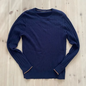 Mörkblå stickad tröja Jack & Jones L - Säljer en mörkblå stickad tröja från Jack & Jones Premium i storlek L. Tröjan har ett snyggt kabelstickat mönster, rund halsringning och diskreta orange detaljer vid ärmsluten. Perfekt för dig som gillar klassisk och stilren look.