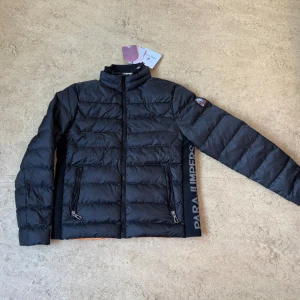 Svart dunjacka från Parajumpers - Snygg svart dunjacka från Parajumpers med orange foder och logga på ärmen. Jackan har dragkedja framtill, två dragkedjefickor och Parajumpers-tryck längs sidan. Perfekt för kalla dagar och har en modern, sportig look.