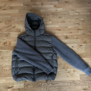 Grå pufferjacka med stickade ärmar - Säljer en mörkgrön pufferjacka från Massimo dutti med stickade ärmar och huva. Jackan har dragkedja framtill, två sidofickor med dragkedja och justerbar dragsko nertill. Perfekt för kalla dagar när du vill ha både stil och värme. Köptes ny på Mos och är i nytt skick, priser ligger därför på 2000 men kan diskuteras. 