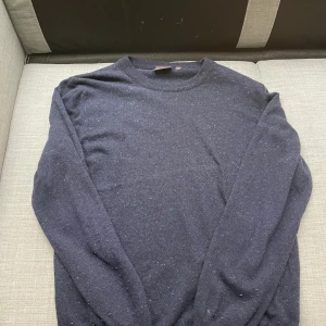 Mörkblå stickad tröja Oscar Jacobson - Mysig mörkblå stickad tröja från Oscar Jacobson, tillverkad i Italien. Tröjan har diskreta färgprickar i garnet och rund halsringning. Perfekt för kyligare dagar och enkel att matcha med olika outfits.