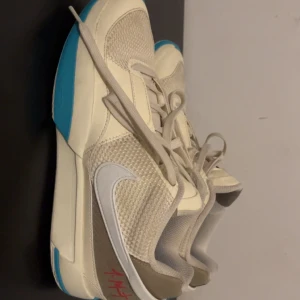 Nike Ja Morant sneakers beige/blå - Säljer ett par Nike Ja Morant sneakers i beige och vitt med blå detaljer på sulan. Skorna har meshpaneler för extra ventilation, klassisk snörning och en vit swoosh på sidan. Snyggt röd Ja Morant-signatur på hälen och mönstrad yttersula med rött logotryck. Lite använd men funkar ändå 😁