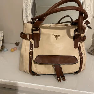 Beige och brun handväska från Poaba - Stilren handväska i beige och brunt från Poaba med snygga spänndetaljer och justerbar axelrem. Väskan har flera fack, dragkedja på baksidan och dekorativa gulddetaljer framtill. Tillverkad i syntetmaterial och rymmer allt du behöver till vardagen.