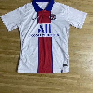 PSG vit matchtröja Nike herr - Paris Saint-Germain fotbollströja i vitt med bred röd och blå rand mitt på, blå krage och klubbmärke på bröstet. Nike-logga och sponsortryck framtill. Tillverkad i lätt och ventilerande material, perfekt för match eller träning.