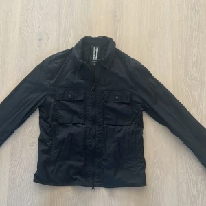 Svart vindjacka från Bomboogie, S - Svart vindjacka från Bomboogie i storlek S. Jackan har två stora bröstfickor med knappar, dragkedja framtill och klassisk krage. Materialet är vattenavvisande, perfekt för blåsiga dagar. Enkel och stilren design som funkar till det mesta. Nypris ca 2700kr | Pris kan diskuteras!