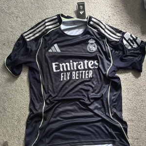 Real Madrid bortatröja Adidas L - Snygg Real Madrid bortatröja från Adidas i mörkblått med tunna ränder, silverfärgade detaljer och klubbmärke på bröstet. Tröjan har korta ärmar, tre klassiska Adidas-ränder på axlarna och är tillverkad i lätt funktionsmaterial.