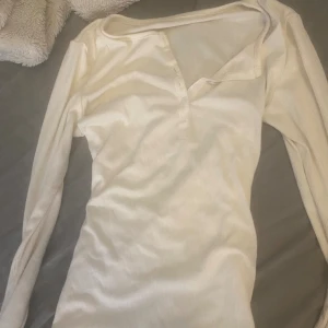 Vit ribbad långärmad topp med knappar - En stilren vit långärmad topp med ribbad struktur och knappar framtill. Toppen har en smickrande v-ringning och är tillverkad i ett mjukt material som känns skönt mot huden. Perfekt att matcha med jeans eller kjol för en enkel och snygg look.