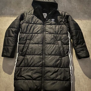 Svart lång pufferjacka från Adidas - Lång svart pufferjacka från Adidas med huva och dragkedja framtill. Jackan har de klassiska vita Adidas-ränderna längs sidorna och logga på bröstet. Quiltad design och två snedställda fickor framtill. Perfekt för kalla dagar. Hör av vid minsta fundering 💫