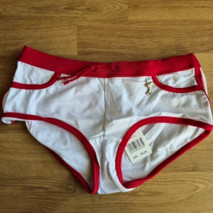 Hipster trosa med Snobben på  - Supergulliga vita hipstertrosor med röda kanter och snörning i midjan. Framtill finns en liten Snoopy-charm och baktill ett tryck med Snoopy i rött. Mjukt material i 95% bomull och 5% elastan för skön passform. Perfekt för dig som gillar söta detaljer och tecknade motiv. Är aldrig använd, kommer från ett konkurslager. Se även mina andra auktioner. Bundlerabatt på 20% vid flerköp. 