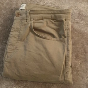 Beige chinos från These Glory Days i storlek 52R - Snygga beige chinos från These Glory Days med klassisk rak passform och normal midja. Byxorna har fickor fram och bak, stängs med knapp och dragkedja. Tillverkade i mjuk bomull som är skön att ha på sig hela dagen.