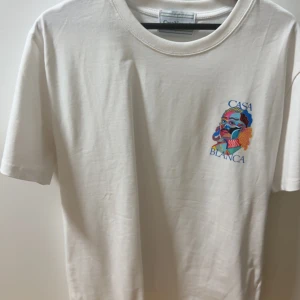Vit t-shirt från Casablanca med tryck - Vit t-shirt från Casablanca med färgglatt grafiskt tryck och texten 'CASABLANCA' både fram och bak. Klassisk passform med rund hals och korta ärmar. Perfekt för dig som gillar streetwear och snygga prints. Oanvänd 