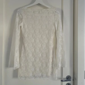 Vit spetsklänning med öppen rygg - Superfin vit kort spetsklänning med långärmade ärmar och öppen rygg som knyts med ett band i nacken. Klänningen har blommigt spetsmönster och lätt utsvängda ärmar. Endast använd en gång! Jätte lite smink på insidan av kragen