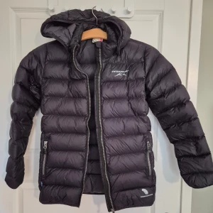 Svart dunjacka från Swedemount - Svart dunjacka från Swedemount med huva och två sidofickor med dragkedja. Jackan har ett quiltat mönster och logotyp på bröstet och ryggen. Perfekt för kalla dagar med sin fluffiga och värmande design. Långärmad och med hel dragkedja framtill.