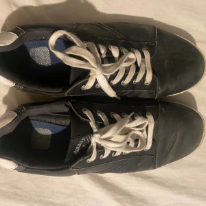 Svarta Callaway sneakers med blå sula - Svarta sneakers från Callaway med vit snörning och vit detalj på hälen. Snygg blå och transparent sula med Callaway-logga och mönstrad undersida. Skorna har rund tå och platt sula, tillverkade i skinn för en klassisk look.