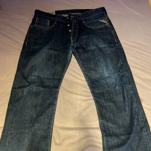 Replay Blue Jeans mörkblå 33/32 - Mörkblå jeans från Replay med klassisk femficksdesign, kontrastsömmar och knappgylf. Modellen har raka ben och snygga detaljer på bakfickorna. Tillverkade i slitstarkt denim med en cool vintagekänsla. Perfekt för dig som gillar streetwear.