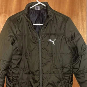 Mörkgrön pufferjacka från Puma XL - Snygg mörkgrön pufferjacka från Puma i storlek XL(barn storlek). Jackan har klassisk quiltad design, dragkedja framtill och två sidofickor. Puma-logga på bröstet och ryggen. Perfekt för kyliga dagar och enkel att matcha med streetwear.