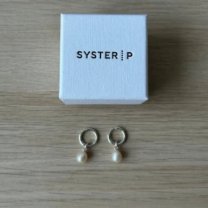 Syster P silverörhängen med pärlor - Snygga örhängen från Syster P i silver med droppformade vita pärlor. Aldrig använda, nypris 800kr. Runda hoops med klassisk design och elegant pärldetalj som ger en stilren vibe. Perfekta för dig som gillar minimalistiska smycken med en twist.