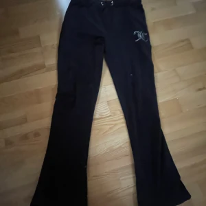 Svarta mjukisbyxor Juicy Couture - Svarta mjukisbyxor från Juicy Couture med bootcut-passform och snörning i midjan. Byxorna har en liten glittrig JC-logga framtill och är tillverkade i mjukt material som känns riktigt skönt mot huden.