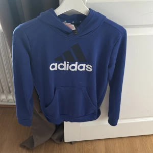 Blå Adidas hoodie med logga - Snygg blå hoodie från Adidas med stor vit och svart logga på bröstet. Tröjan har huva, känguruficka och långa ärmar. Perfekt för dig som gillar sportig och avslappnad stil. Det vita är inte fläck det är kameran. 