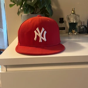 Röd baseboll NY Yankees keps - Säljer en röd baseboll keps med New York Yankees-logga broderad i vitt framtill. Klassisk flat brim och broderade ventilationshål. Kepsen har New Era-logga på sidan och MLB-logga bak. Perfekt för dig som vill sticka ut med en sportig streetstyle.