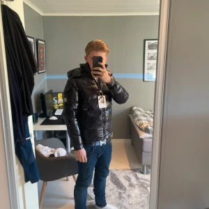 Svart Moncler pufferjacka med huva - Säljer en svart glansig pufferjacka från Moncler med huva och dragkedja. Jackan har en quiltad design och klassisk Moncler-logga på ärmen. Perfekt för kalla dagar och riktigt snygg till jeans. Sitter normal på kroppen och har långa ärmar.