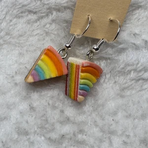 Regnbågsörhängen i form av tårtbitar - Superfina örhängen formade som små tårtbitar med regnbågsfärger. De har bågar i silverfärg och detaljer i lila, blått, grönt, gult, orange och rött. Perfekta för dig som vill sticka ut med färgglada accessoarer.
