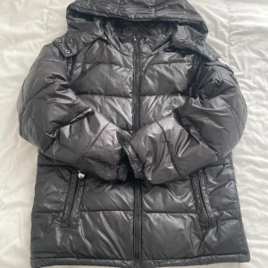 Svart pufferjacka med huva falsk moncler  - Säljer en svart pufferjacka med huva och dragkedja framtill. Jackan har två fickor med dragkedja och quiltad design. Perfekt för kalla dagar och har en snygg, lite glansig finish. Passformen är normal och jackan är långärmad.