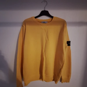 Gul Stone Island sweatshirt L - Gul sweatshirt från Stone Island i storlek L. Klassisk rund halsringning, ribbade muddar och ikonisk logga på vänster ärm. Mjuk bomullskvalitet och relaxed fit gör den perfekt för chill dagar. Snygg och enkel att matcha med jeans eller trackpants.