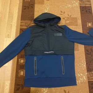 Blå och svart vindjacka Under Armour - Snygg blå och svart vindjacka från Under Armour med huva och dragkedja framtill. Jackan har två praktiska fickor med dragkedja och logga på bröstet. Perfekt för dig som gillar sportig stil och vill ha något lätt och funktionellt.
