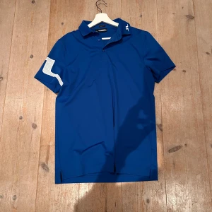 Blå pikétröja från J.Lindeberg - Snygg blå pikétröja från J.Lindeberg med vit logga på kragen och coolt vitt mönster på ärmen. Tröjan har klassisk krage, knäppning framtill och är tillverkad i ett mjukt och stretchigt polyestermaterial. Perfekt för en sportig look.