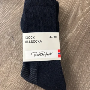 Tjocka svarta ullsockor Pierre Robert - Mörka, tjocka ullsockor från Pierre Robert i storlek 37/40. Perfekta för kalla dagar, med en blandning av ull, polyester och polyamid för extra värme och komfort. Diskret design utan mönster, passar dig som gillar klassisk stil.