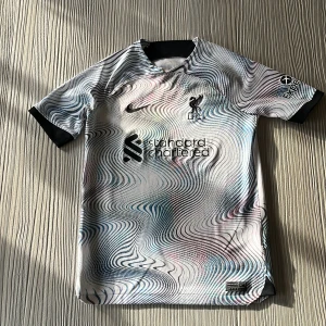 Liverpool Nike Dri-Fit fotbollströja L - Liverpool FC fotbollströja från Nike med Dri-Fit-material. Tröjan har ett unikt vågigt mönster i svart, blått och grönt på vit bas, broderad LFC-logga och sponsortryck framtill. Svarta detaljer vid krage och ärmslut. Perfekt för dig som älskar fotboll och vill sticka ut. Den är äkta köpt från Nike. Storlek 147-158