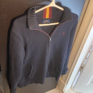 Mörkblå stickad tröja Polo Ralph Lauren - Mörkblå stickad tröja från Polo Ralph Lauren med dragkedja framtill och ribbad krage med röda och gula ränder. Klassisk röd logga broderad på bröstet och två fickor framtill. Perfekt för kyliga dagar och snygg till jeans. Nypris 1500, mitt pris 500. Passar mellan 180-185