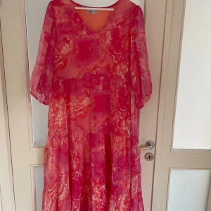Rosa långklänning från Isolde XL - Säljer en flowy långklänning från Isolde i storlek XL. Klänningen har ett coolt abstrakt mönster i olika rosa och koralltoner, v-ringad hals och halvtransparenta, luftiga ärmar. Perfekt för dig som gillar färg och vill sticka ut med en unik look.